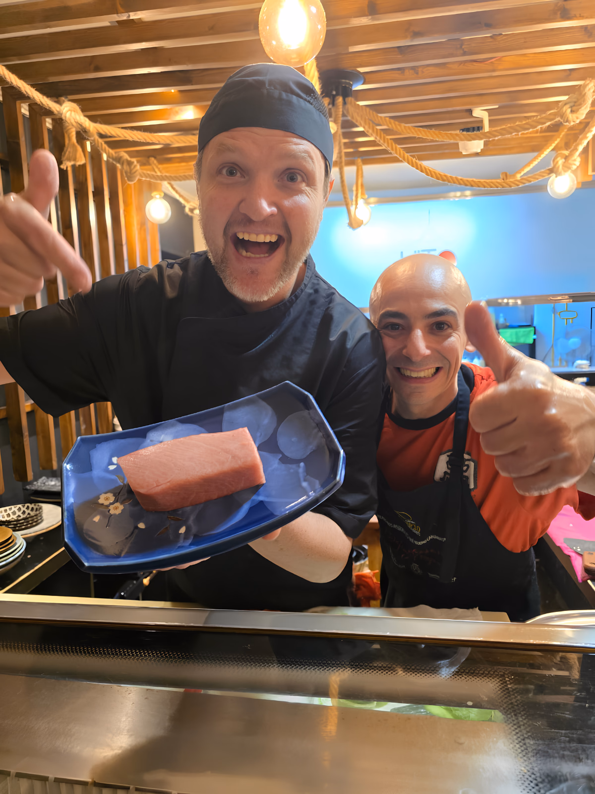 Oliver Schultz sosteniendo un lomo entero de atún rojo Balfegó en un plato azul en la barra de Hito, junto a un compañero que levanta el pulgar