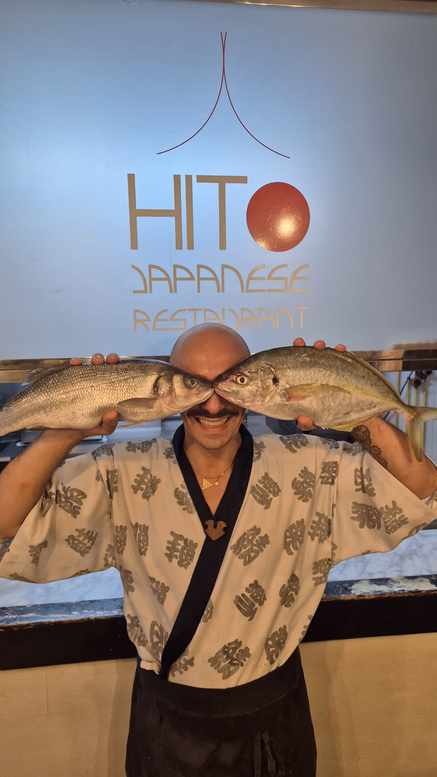 El chef de Hito sosteniendo dos pescados frescos frente al logo del restaurante